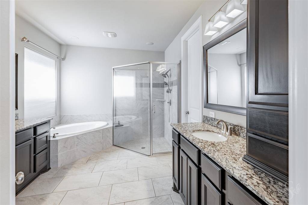 The spa-inspired primary bath features dual granite topped vanities, a deep soaking tub, and an oversized glass-enclosed shower with elegant tilework. Rich cabinetry and upscale lighting complete this luxurious space designed for relaxation.