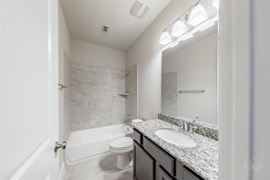 The first-floor guest bath is beautifully finished with a granite countertop, rich cabinetry, and oversized tile flooring. A tile-surround tub/shower combo and modern lighting add a touch of upscale refinement to this elegant space.