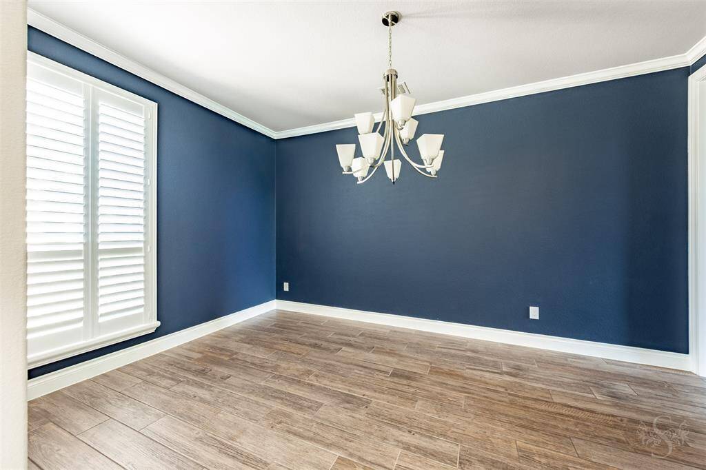 The formal dining room exudes sophistication with custom accent walls, crisp white trim, and crown molding under a lovely chandelier. Plantation shutters and wide-plank tile flooring complete this polished space, perfect for upscale entertaining.