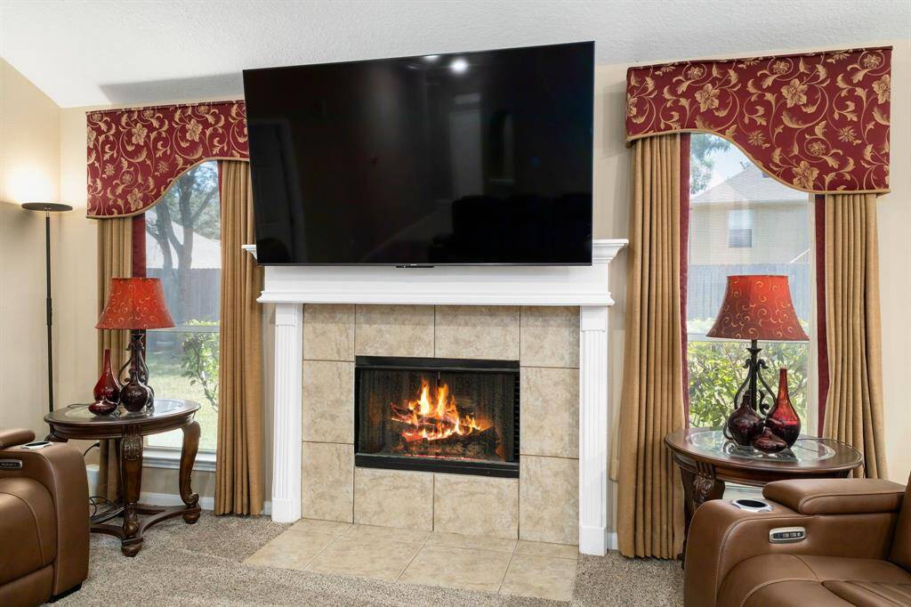 Cozy living room with a tiled fireplace and wood mantle surrounding.  Features large windows and a welcoming atmosphere.