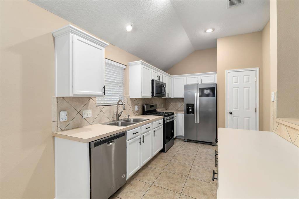 This kitchen features modern stainless steel appliances, white cabinetry, and a neutral tile backsplash. The space is bright with a high ceiling and recessed lighting, offering a clean and functional layout.