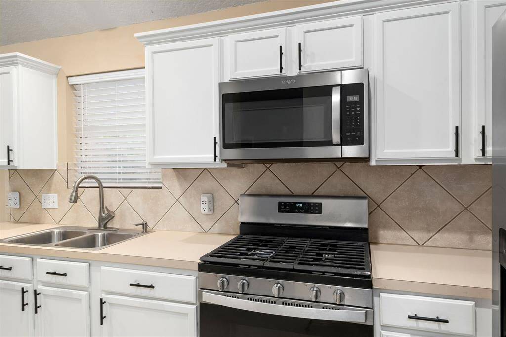 This kitchen features modern stainless steel appliances, including a gas stove and microwave. It has white cabinetry, a double sink, and a neutral tile backsplash, offering a clean and contemporary look.