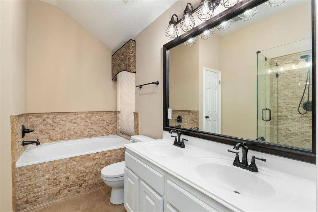 This bathroom features a double vanity with modern fixtures, a large mirror, and a glass-enclosed shower. The space includes a soaking tub with tile surround and neutral tones for a warm, inviting feel.