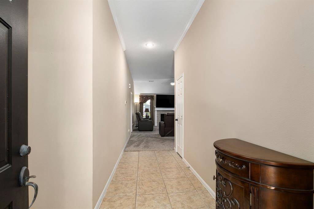 This photo shows a welcoming entry hallway with tiled flooring and neutral walls. It leads to a cozy living area visible in the distance, featuring a carpeted section and a glimpse of a fireplace and seating, creating a warm and inviting atmosphere.