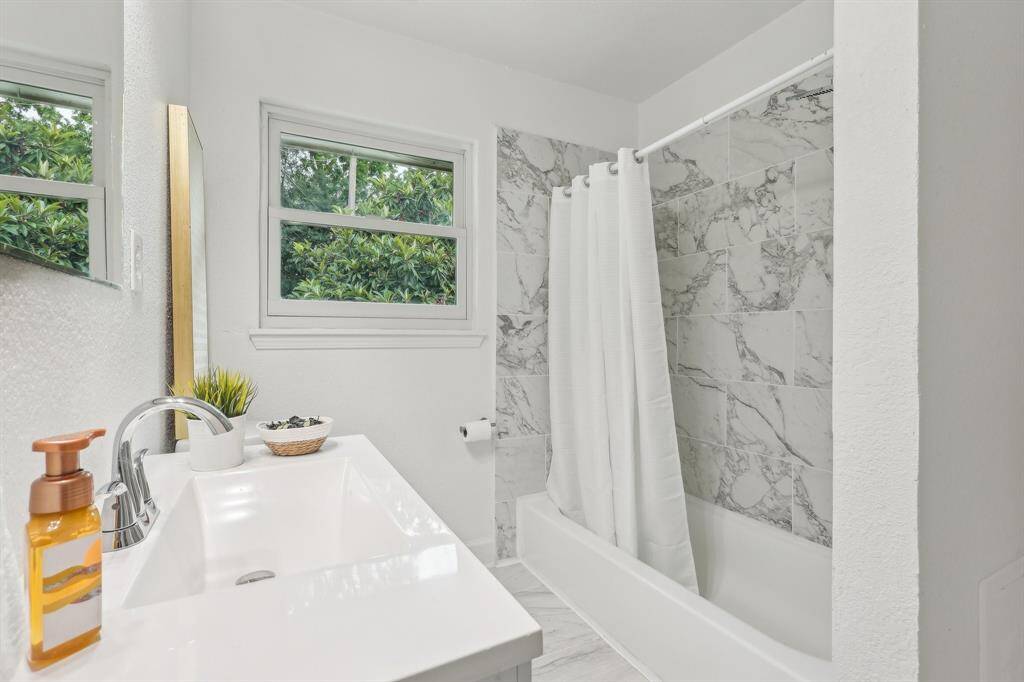 This bathroom features a clean, modern design with a white vanity, chrome fixtures, and a marble-tiled shower. Natural light streams in through two windows, creating a bright and airy atmosphere.