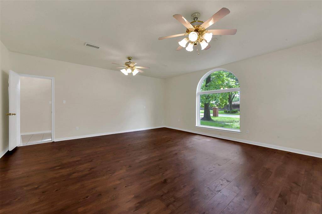 Spacious room with hardwood floors, neutral walls, and large arched window providing natural light. Features two ceiling fans and an open doorway leading to another area.