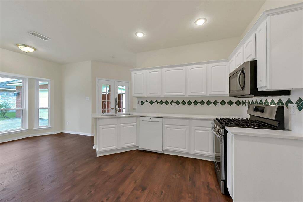 This kitchen features modern stainless steel appliances, white cabinetry, and a decorative tile backsplash. It has an open layout with wood flooring and ample natural light from large windows.