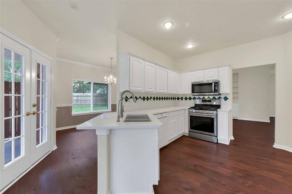 This photo showcases a bright kitchen with white cabinetry and stainless steel appliances. The open layout features dark wood flooring, a modern chandelier, and French doors leading to a backyard view, ideal for natural light and entertaining.