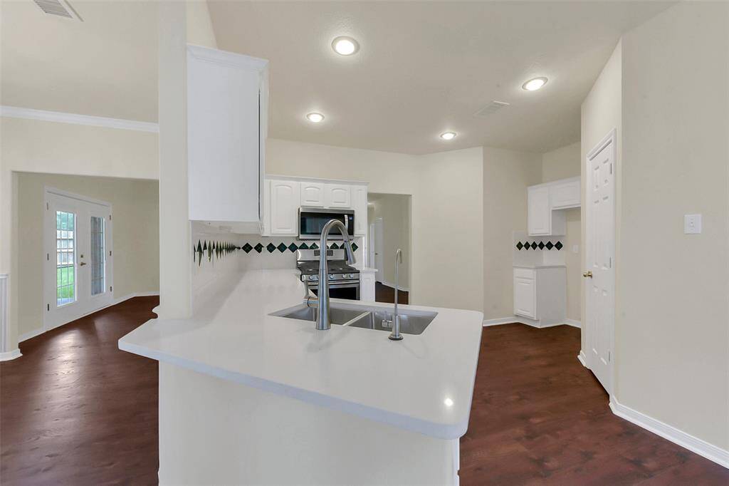 This kitchen features a modern, open layout with a sleek white countertop and stainless steel appliances. It has ample lighting and a view into the adjoining room through a doorway, offering a spacious feel.
