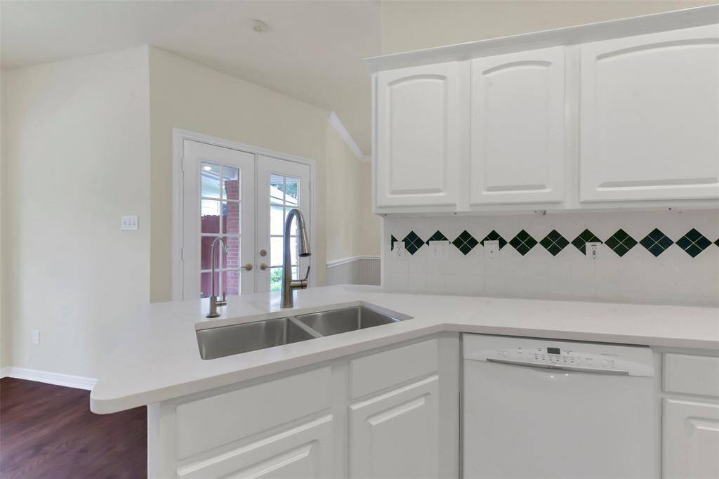 This kitchen features a modern design with white cabinetry, a dual sink, and a dishwasher. The backsplash has a stylish diamond pattern, and French doors provide ample natural light and access to the outdoors.