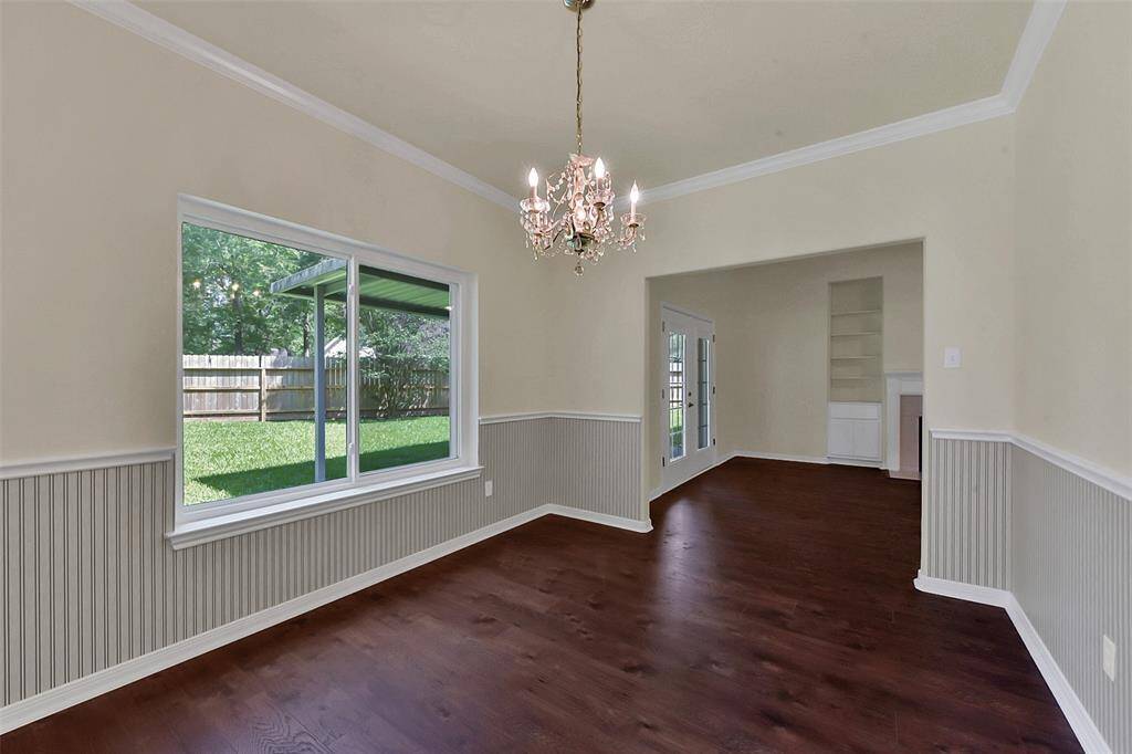 This room features dark hardwood flooring and wainscoting, with a large window overlooking a fenced backyard. It includes a chandelier and opens into another area with built-in shelving and glass doors.