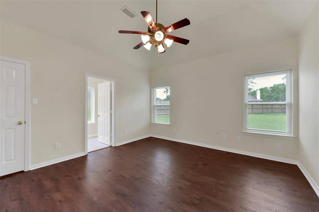This spacious bedroom features hardwood floors, neutral walls, and ample natural light from two windows. A ceiling fan adds comfort, and a doorway leads to an ensuite bathroom.