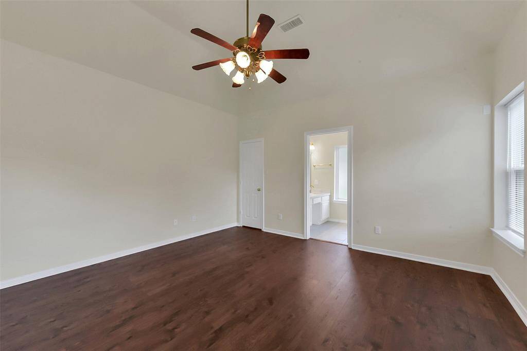 This spacious bedroom features hardwood floors, neutral-colored walls, a ceiling fan, and a large window for natural light. It connects to a bright en-suite bathroom.