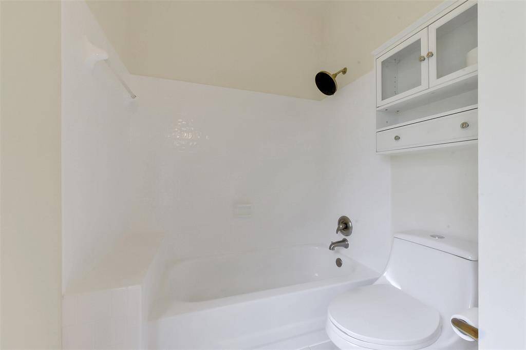 This bathroom features a minimalist design with a white bathtub and shower combination, modern fixtures, and a toilet. A compact storage cabinet is mounted above the toilet, providing convenient space for essentials.