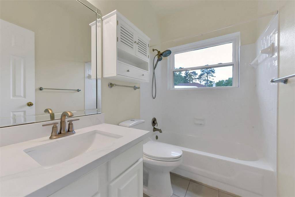 This is a bright, clean bathroom featuring a white vanity with a modern faucet, a bathtub with a shower, and a window providing natural light. The space includes ample storage with a wall cabinet and towel bars.
