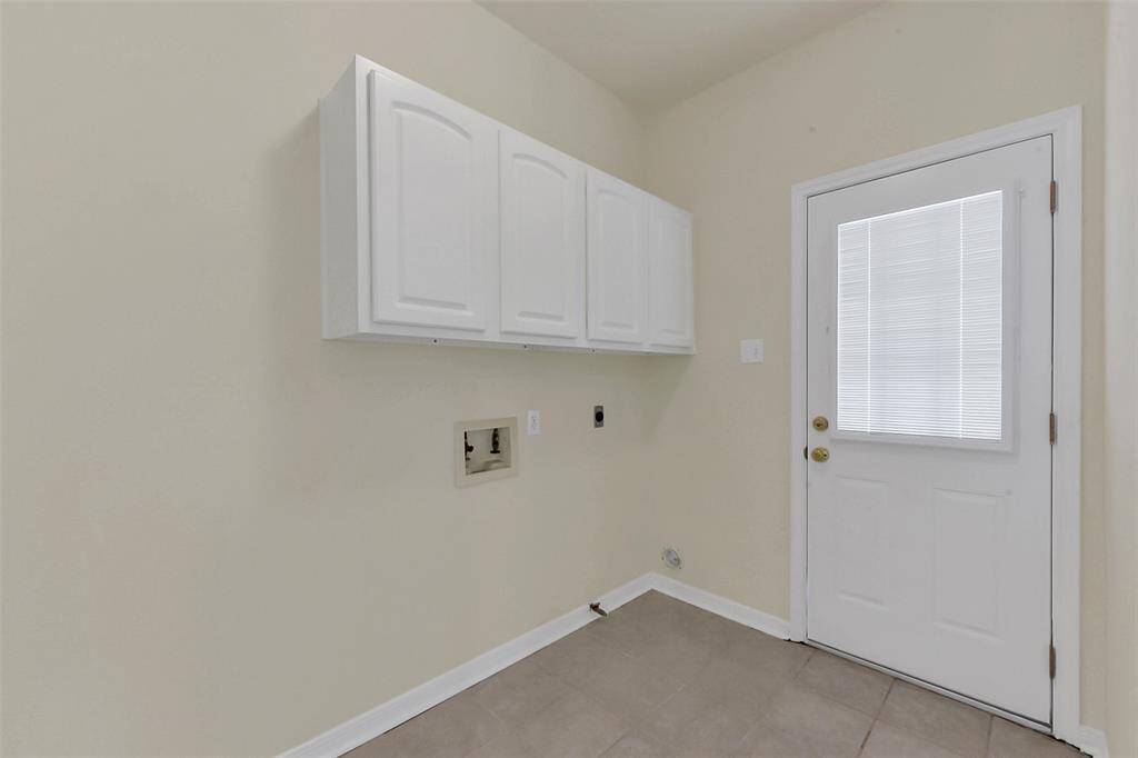 This photo shows a utility room with light-colored walls, tiled flooring, and a white door with a window. There are white cabinets mounted on the wall for storage and hookups for laundry appliances.