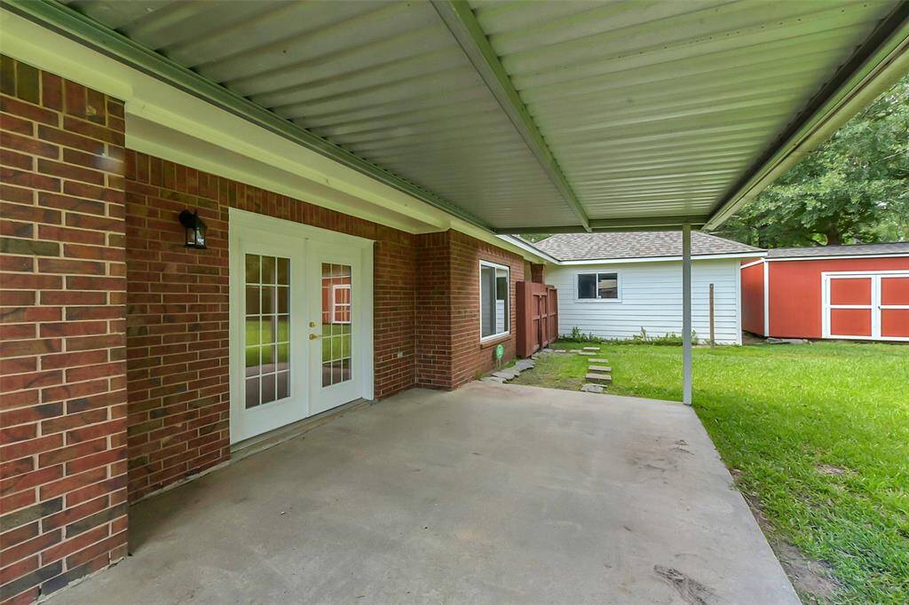 This photo showcases a covered patio area with a concrete floor and brick exterior, featuring glass double doors. It opens to a grassy backyard with a detached structure in the background, offering a versatile outdoor space.