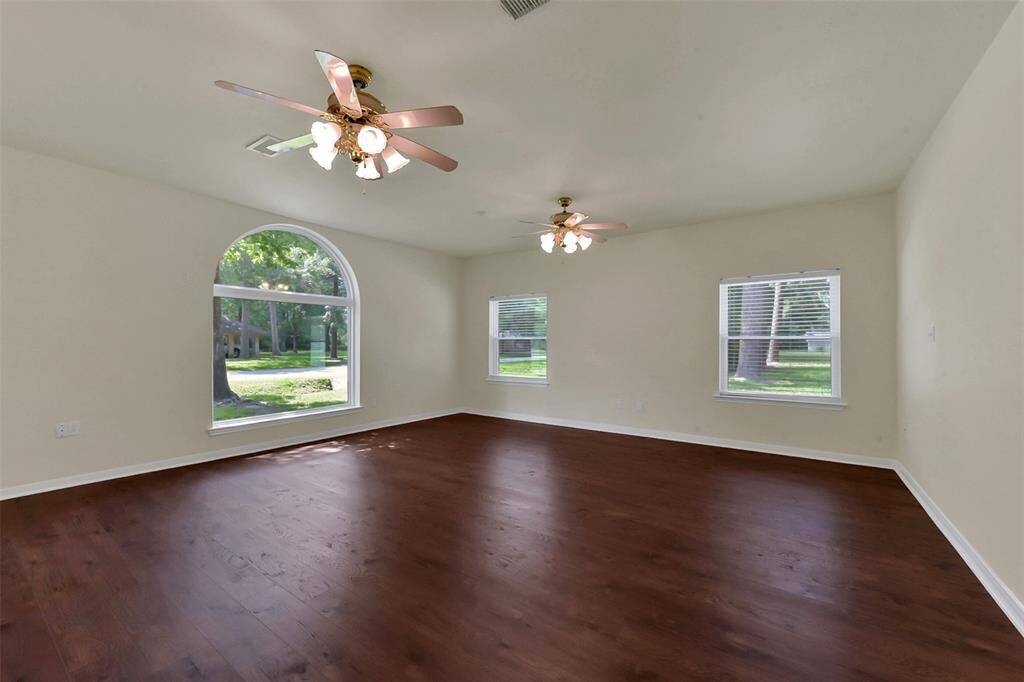 Spacious living room with rich wood floors, neutral walls, and abundant natural light from large windows. Features ceiling fans and a serene view of the greenery outside.