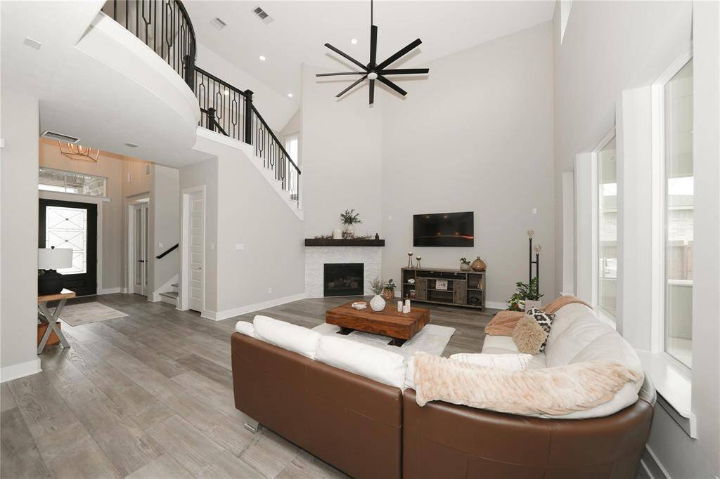 This spacious living room features a high ceiling with a modern fan, a cozy fireplace, and large windows for natural light. The open floor plan includes a stylish staircase and a welcoming entryway, creating an inviting atmosphere perfect for entertaining.