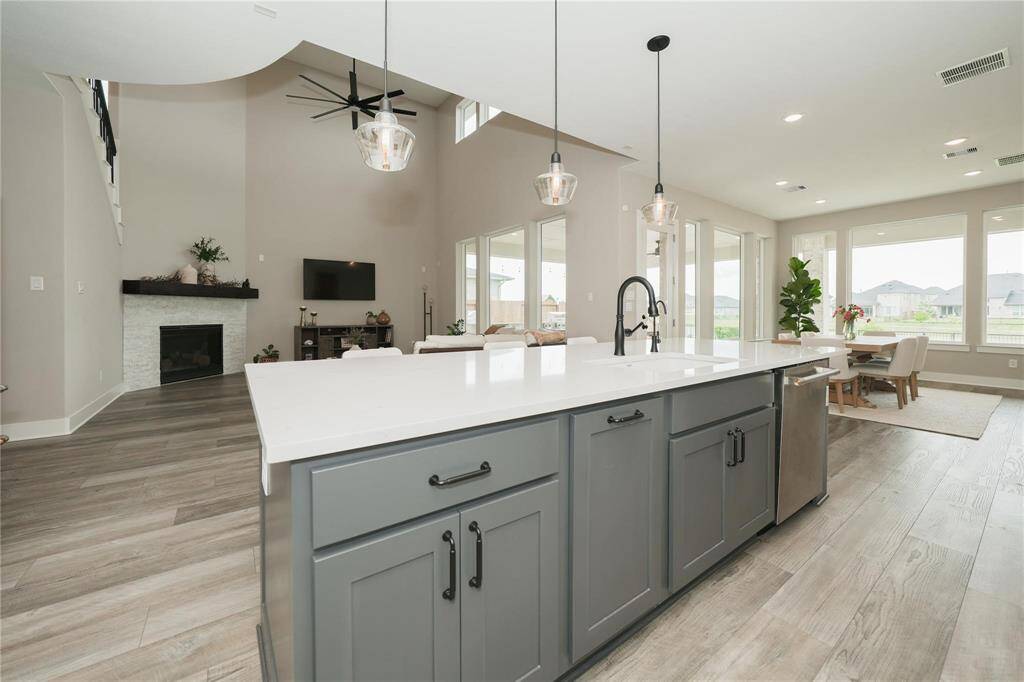 This spacious, open-concept living area features a modern kitchen with a large island and sleek countertops, flowing seamlessly into a bright living room with high ceilings, a ceiling fan, and a fireplace. Expansive windows offer plenty of natural light and views of the outdoors, while the dining area provides a cozy space for meals.