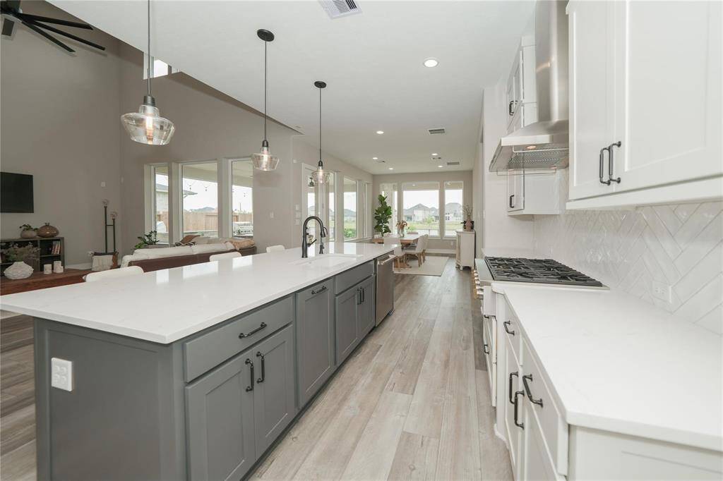 This photo showcases a modern, open-concept kitchen with sleek white cabinetry, a large island with a dark finish, and pendant lighting. The space features stainless steel appliances, a gas stove, and opens into a bright living area with large windows offering plenty of natural light.