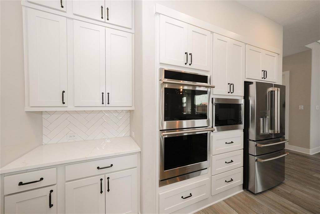 This kitchen features modern stainless steel appliances, including double ovens and a sleek refrigerator. White cabinetry with black handles and a herringbone backsplash add a stylish touch, complemented by light wood flooring.