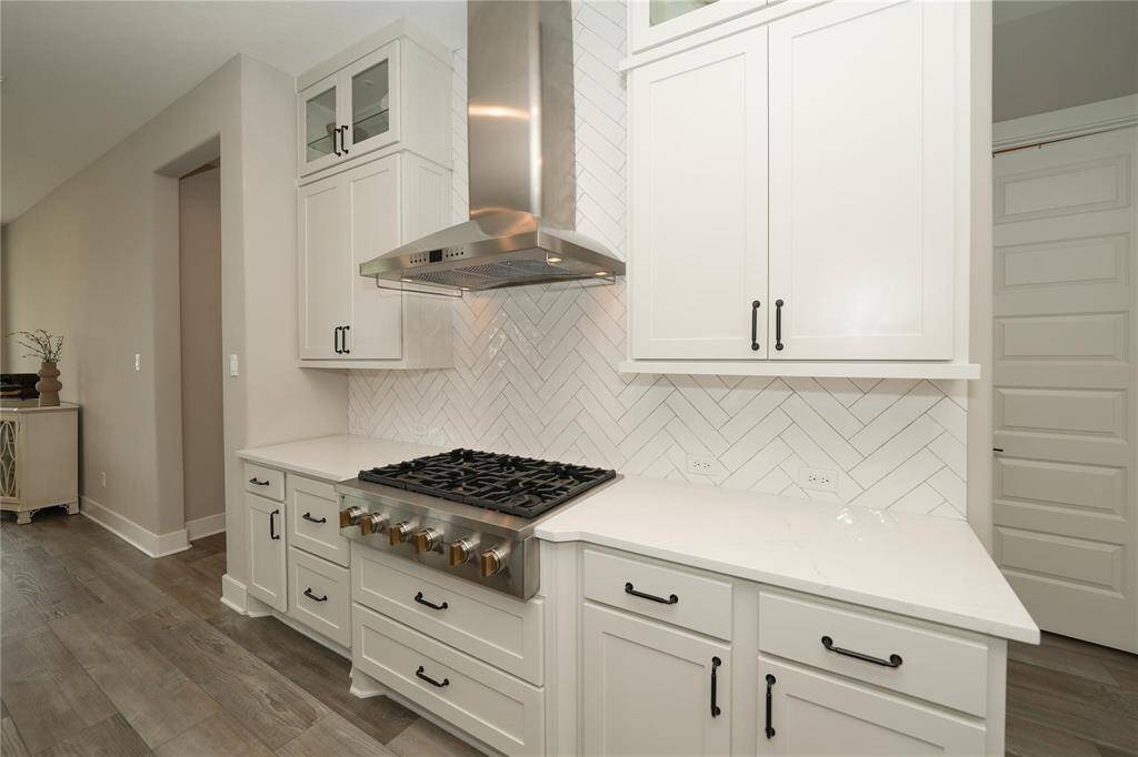 This kitchen features modern white cabinetry with black hardware, a sleek stainless steel range hood, and a gas cooktop. The herringbone-patterned backsplash and light countertops add a touch of elegance, complemented by wood flooring. Ideal for a stylish, functional cooking space.
