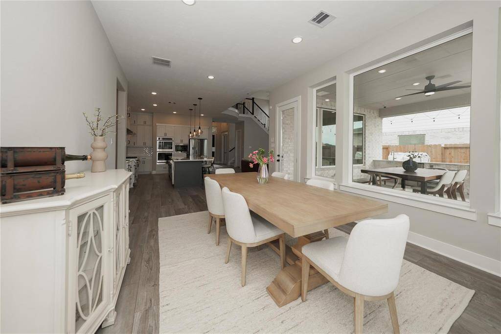 This photo showcases a spacious, modern dining area with a wooden table and white chairs, adjacent to an open kitchen. Large windows provide ample natural light and a view of the outdoor patio. The neutral color palette and stylish decor create an inviting atmosphere.