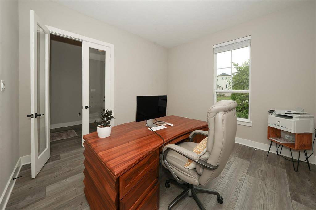 This photo shows a bright, modern home office with a large wooden desk, ergonomic chair, and natural light from a window. It features light-colored walls, wood flooring, and a door with glass panels, creating an inviting workspace.