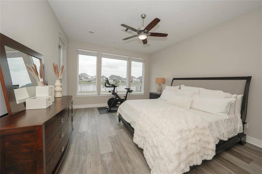 A bright and spacious bedroom with large windows offering scenic views. It features a comfortable bed, stylish dresser, and a ceiling fan. The room includes a stationary bike for fitness enthusiasts and has modern, neutral decor with wood flooring.