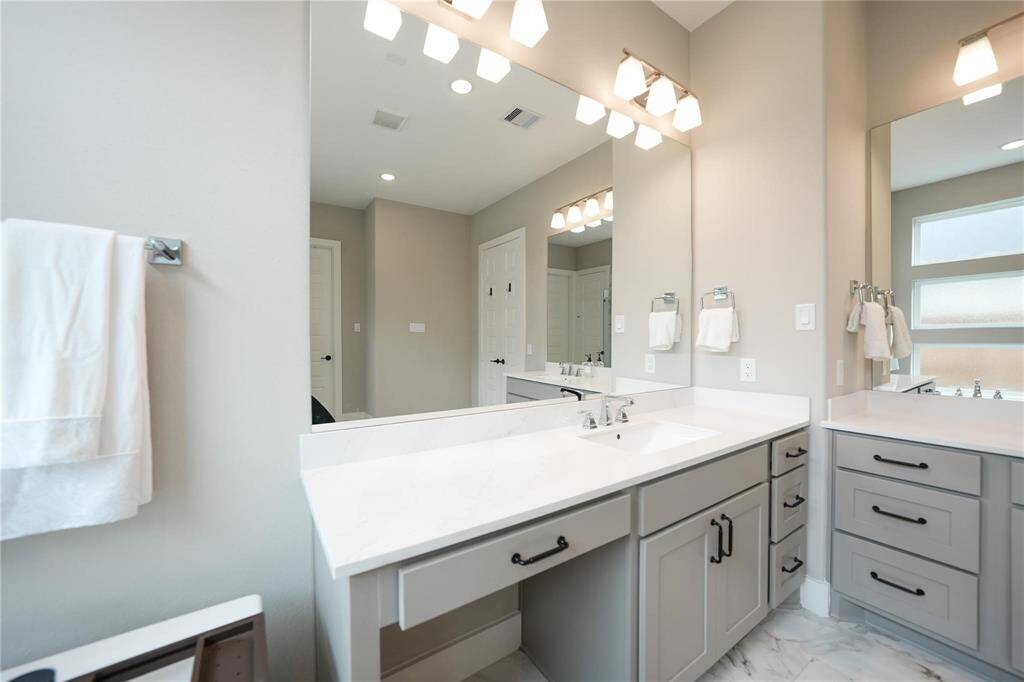 This modern bathroom features a spacious double vanity with sleek white countertops, ample storage, and elegant lighting. The neutral color palette and large mirrors enhance the bright, open feel.