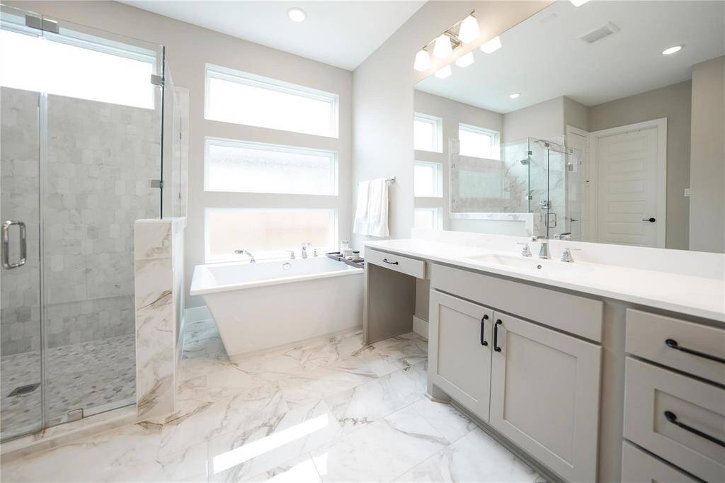 This modern bathroom features a sleek design with a glass-enclosed shower, a freestanding bathtub, and dual sinks. The space is bright with natural light from multiple windows and showcases elegant marble flooring and countertops.
