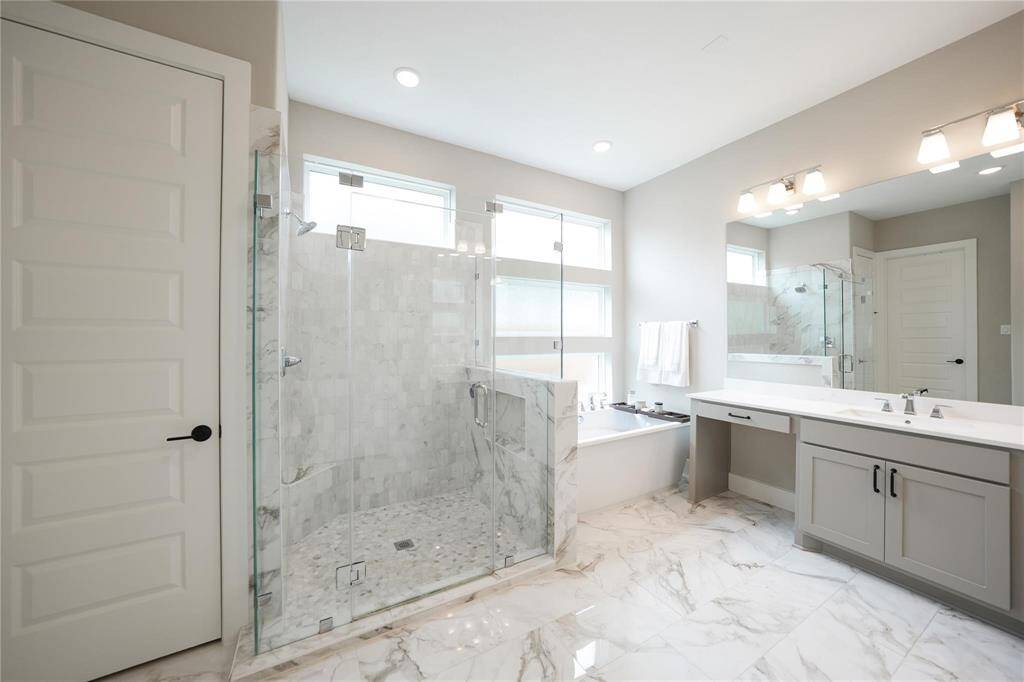 This bathroom features a spacious design with marble flooring and a large glass-enclosed shower. It includes a bathtub next to a double vanity with ample storage, illuminated by modern lighting fixtures. The space is bright and elegant, ideal for a luxurious feel.
