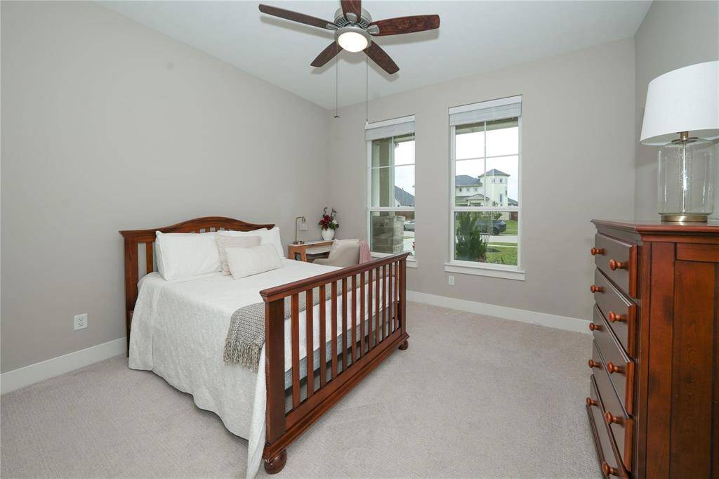 This cozy bedroom features neutral walls and carpet, a wooden bed, a matching dresser, and large windows providing natural light. A ceiling fan adds comfort, and a small desk by the window offers a workspace.