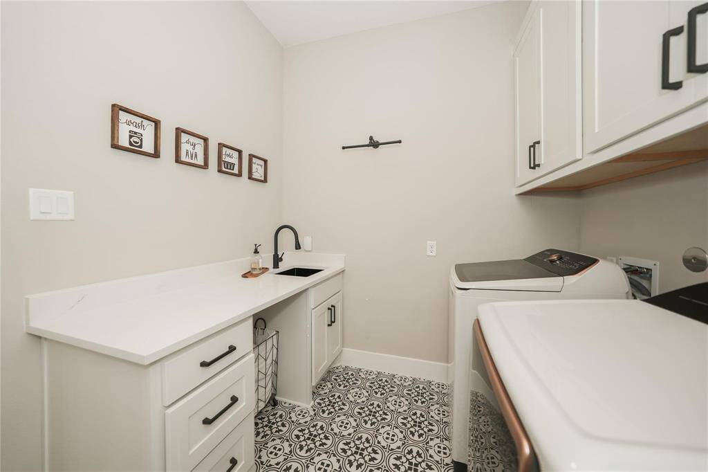 This photo shows a modern laundry room with patterned tile flooring, white cabinetry, and a sleek countertop. It features a washer and dryer, a small sink, and decorative wall art, creating a functional and stylish space.