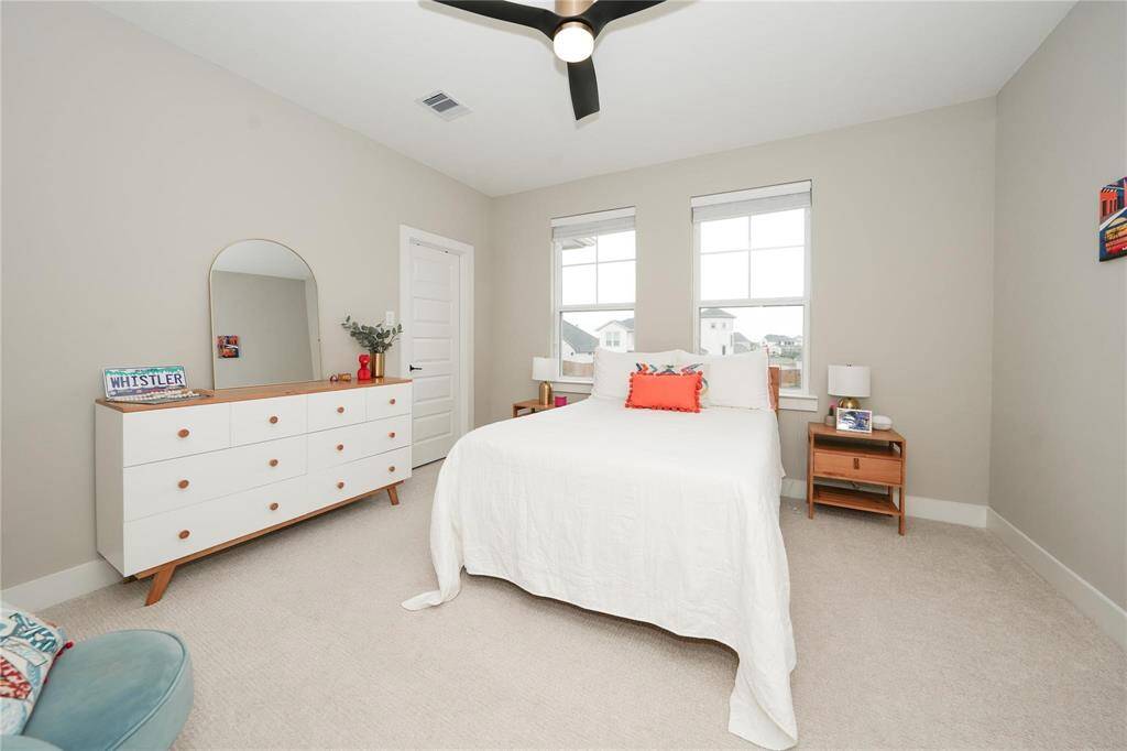 Bright and cozy bedroom with neutral walls and carpet, featuring a double bed, modern ceiling fan, and natural light from two windows. The room includes a stylish dresser and bedside tables for storage.