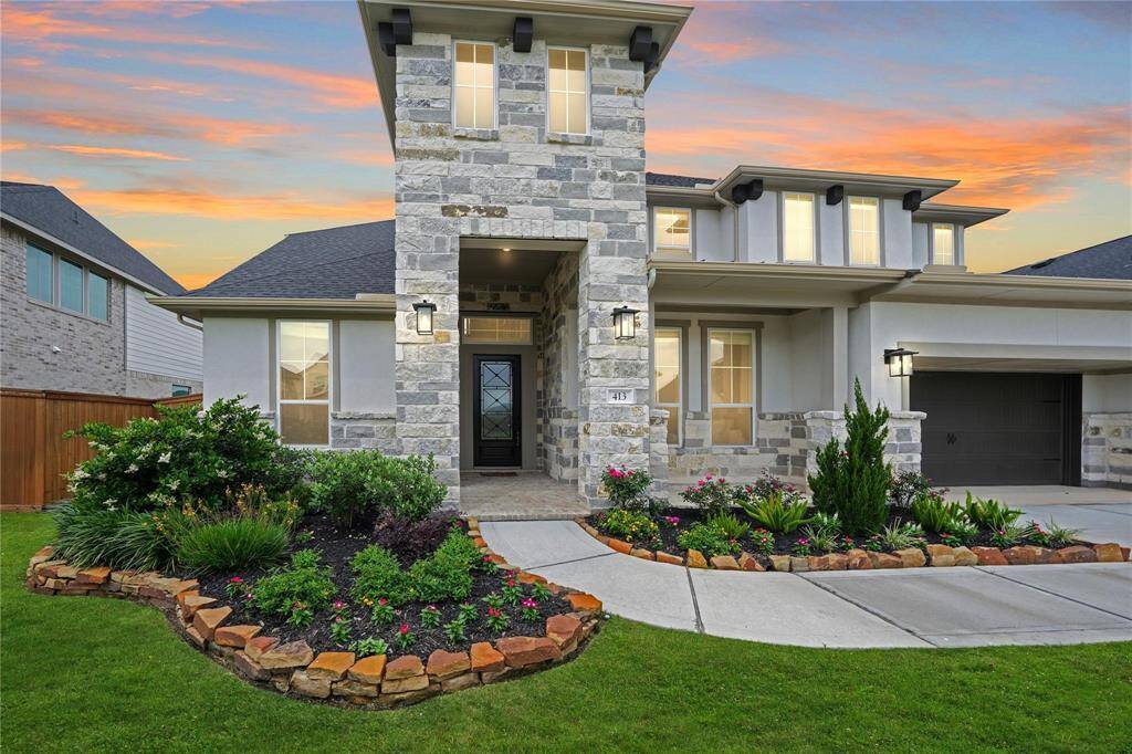 This is a modern two-story stone house with a well-maintained front yard featuring lush landscaping. The entrance is inviting, and the property includes a spacious driveway leading to a garage, all set against a beautiful sunset backdrop.