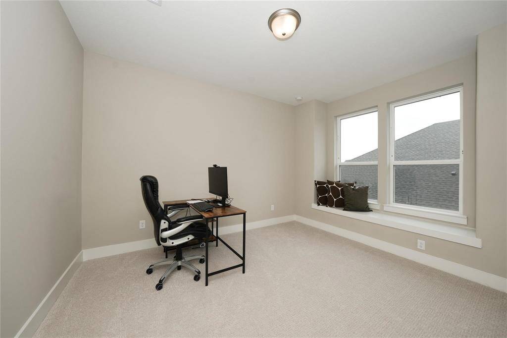 This room features a neutral palette with carpet flooring, a small desk setup, and a cozy window nook with cushions, ideal for a home office or reading area.