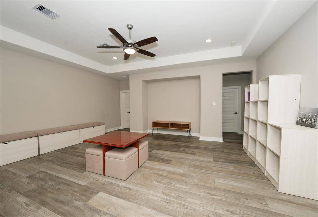 This spacious room features modern wood-look flooring, a ceiling fan, and recessed lighting. It includes built-in storage benches, open shelving, and neutral walls, offering a versatile and inviting space.