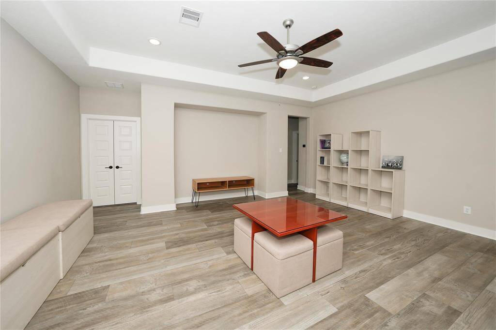 This room features modern wood-look flooring, neutral walls, and recessed lighting. It includes a ceiling fan, built-in shelving, a small bench, and a vibrant red coffee table. Double doors and an open layout provide flexibility for use.