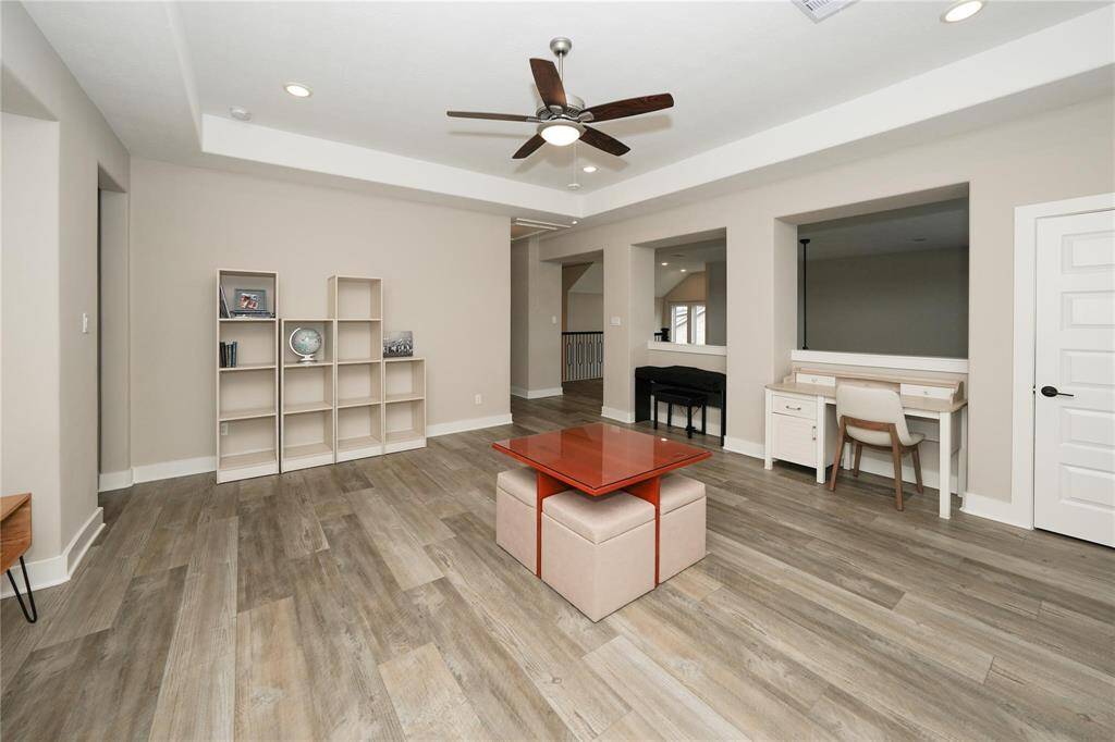 This spacious room features modern wood flooring, a ceiling fan, and recessed lighting. It includes a built-in desk area, open shelving, and a stylish central table with cushioned seating, perfect for a versatile living or workspace.