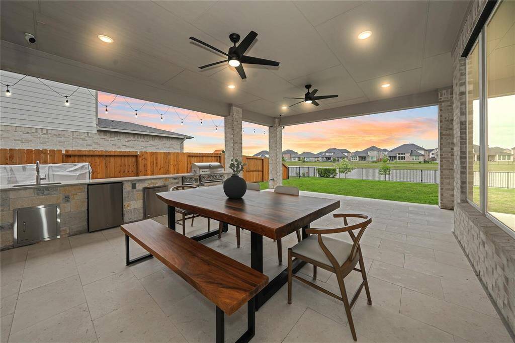This photo showcases a spacious covered patio with modern amenities, including a built-in outdoor kitchen and stylish dining area. The space features ceiling fans, recessed lighting, and a view of a well-maintained backyard with a serene neighborhood backdrop, perfect for entertaining and relaxation.