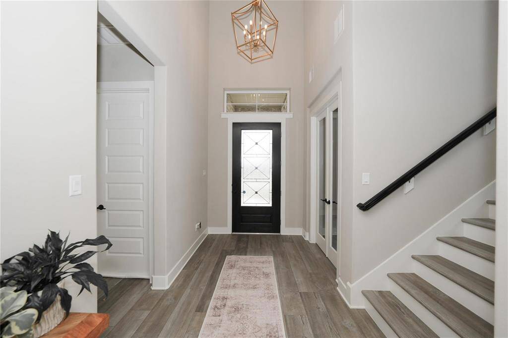 This entryway features a modern design with light wood flooring, a stylish black front door with glass panels, and a geometric pendant light. The space includes a staircase with a sleek black railing and access to adjacent rooms through white doors.