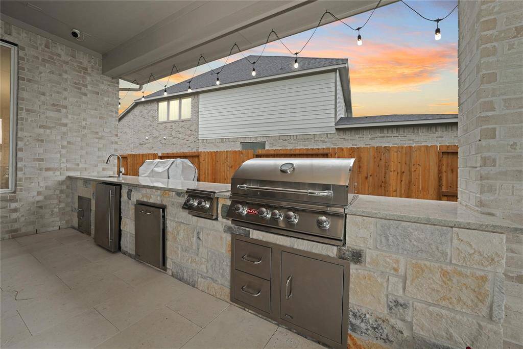 This photo showcases a modern outdoor kitchen with a built-in stainless steel grill, ample counter space, a sink, and under-counter storage, all set against a stone and brick backdrop. String lights add a cozy ambiance, perfect for evening gatherings.