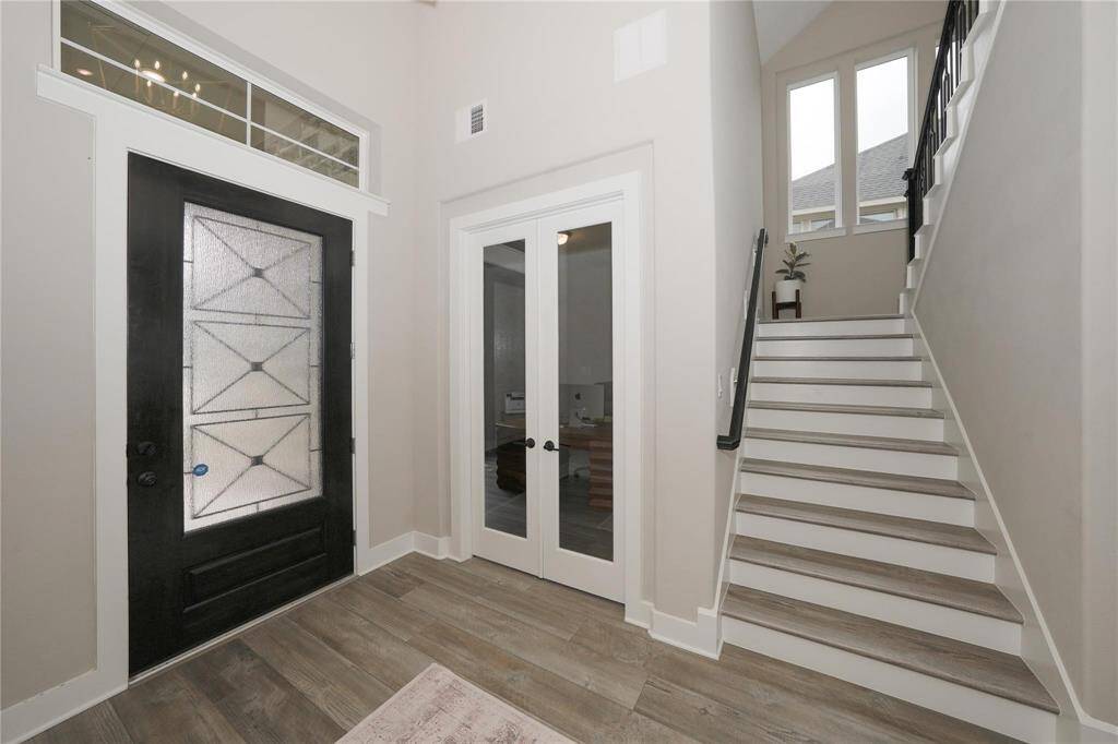 This photo showcases a modern entryway with a stylish black front door featuring frosted glass, a set of French doors leading to another room, and a staircase with black railing. The space is bright and airy with light wood flooring and neutral walls.