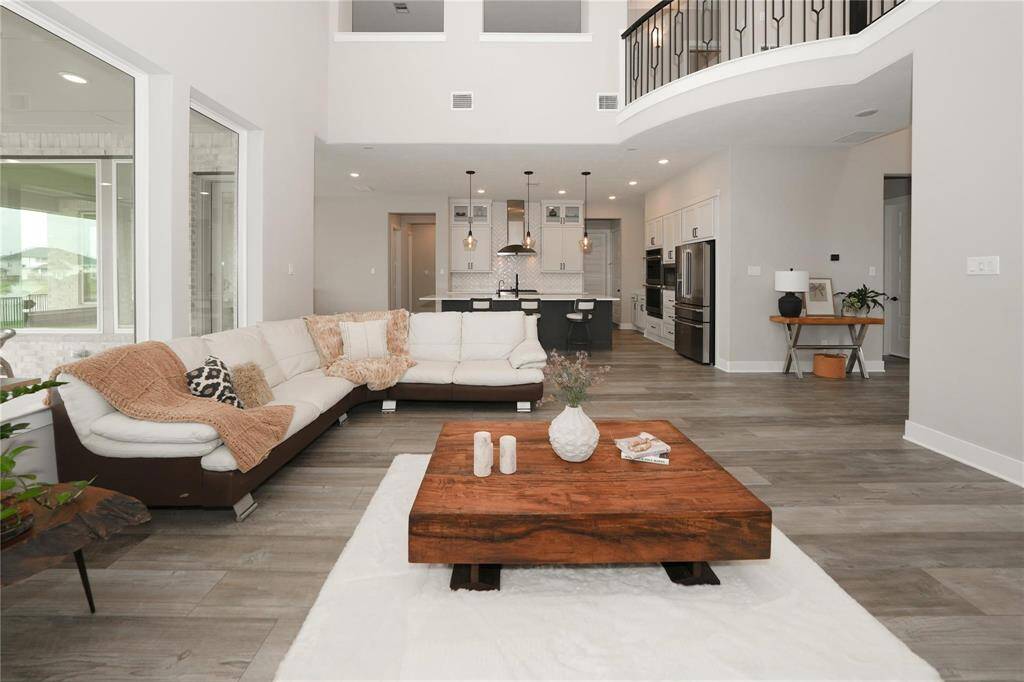 This modern living area features an open floor plan with high ceilings, a cozy sectional sofa, and a central wooden coffee table. The space is brightened by large windows, leading to a sleek kitchen with pendant lighting and stainless steel appliances. Ideal for entertaining and family gatherings.