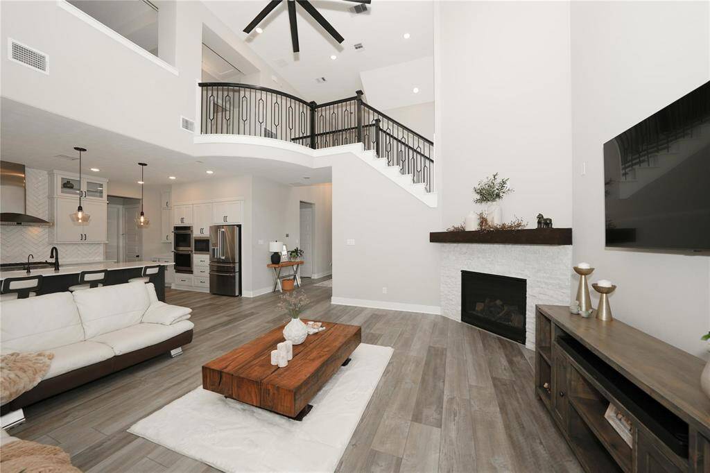 This spacious, modern living room features high ceilings, a sleek ceiling fan, and an elegant staircase with a black railing. The room includes a cozy fireplace and a mounted TV, with an open-concept design leading to a contemporary kitchen equipped with stainless steel appliances. The light wood flooring and neutral color palette create a welcoming atmosphere.