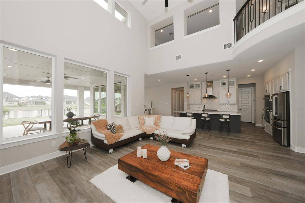 This spacious, modern living room features high ceilings, large windows, and an open floor plan connecting to a sleek kitchen with an island. The room is bright and airy, offering a view of a serene outdoor area with a patio.