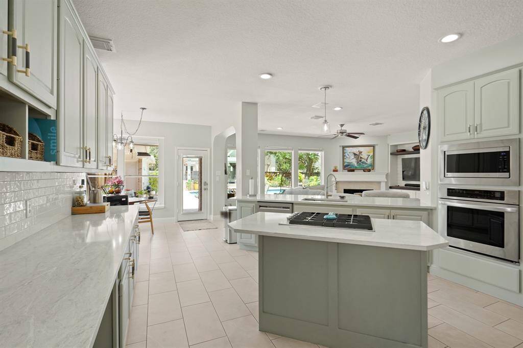 This bright, open-concept kitchen features modern appliances, a central island with a cooktop, and ample counter space. It flows seamlessly into the living area, with large windows offering views of a lush backyard and pool.