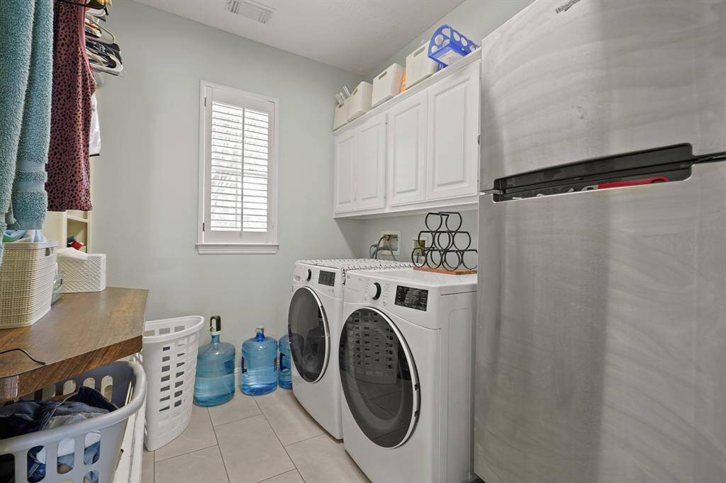 This laundry room features a washer and dryer, ample cabinet storage, a stainless steel fridge, and natural light from a window with blinds.